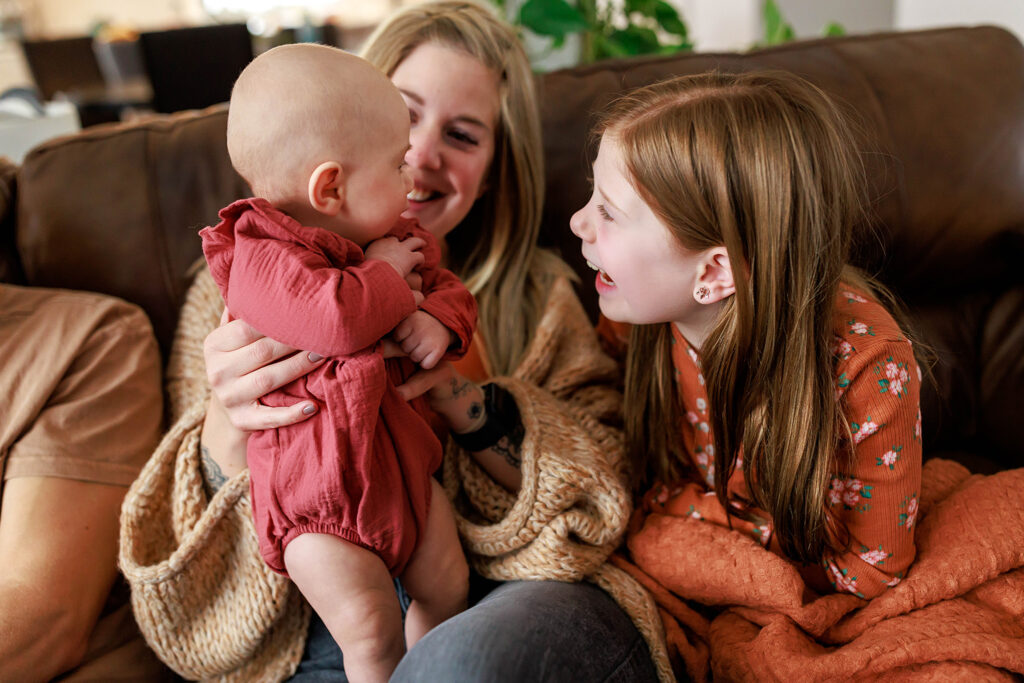 young girl and mom looking at baby sister and smiling during family in home photo shoot