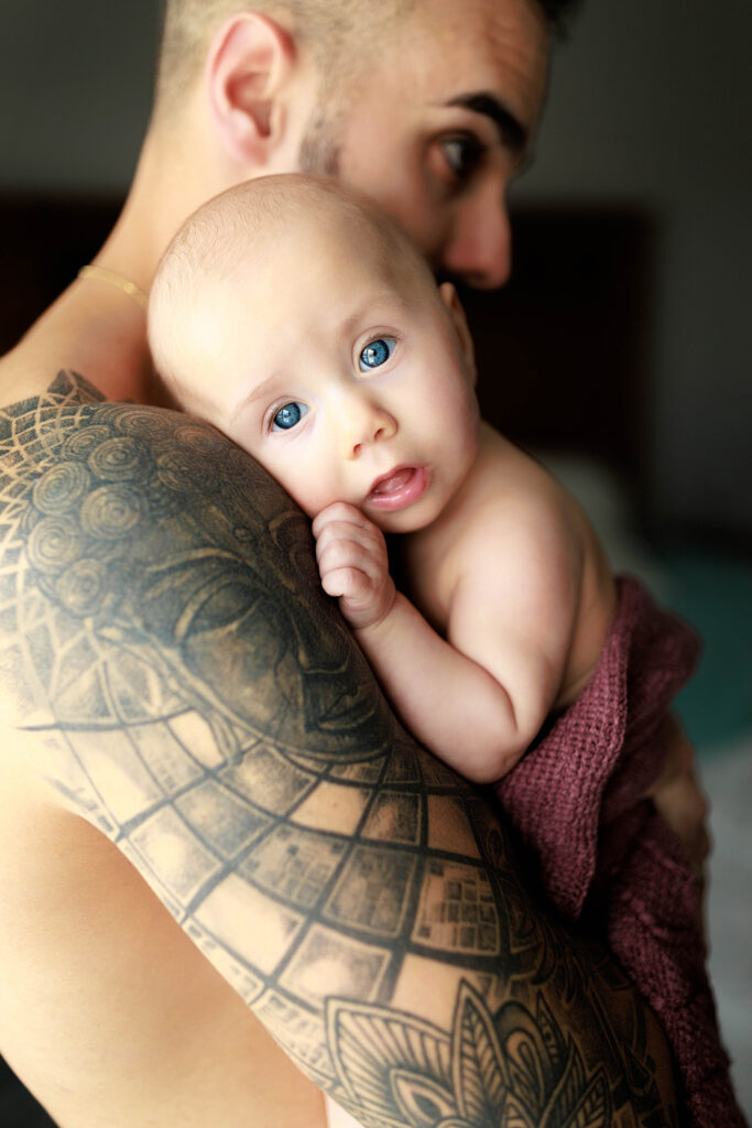 shirtless dad with tattoos holding baby with blue eyes