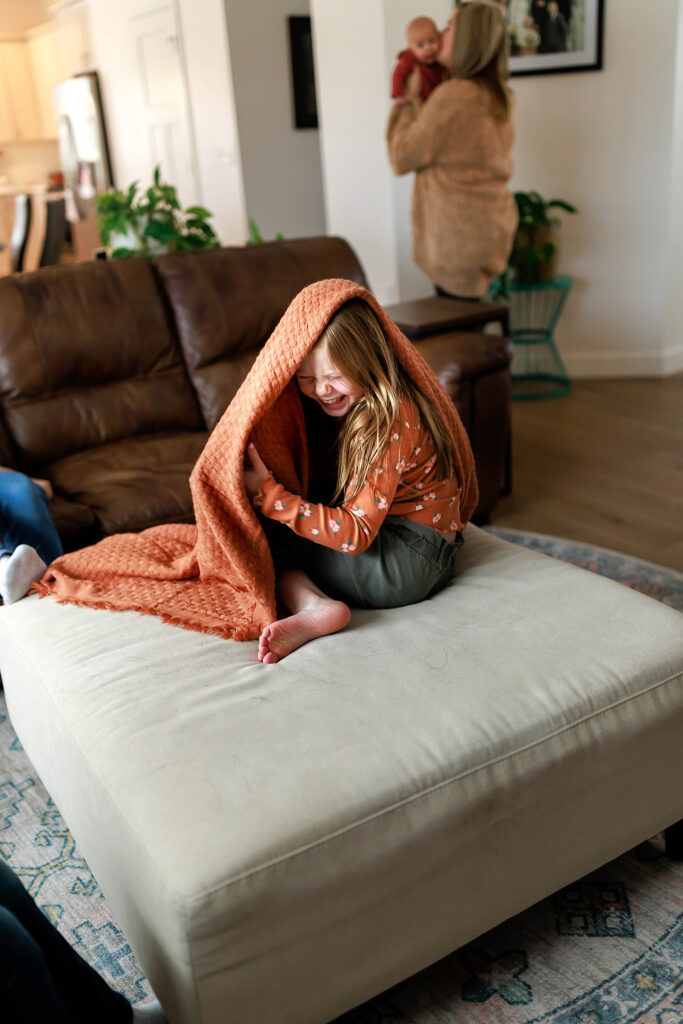 young girl giggling as she hides under blanket in living room during in home photo shoot