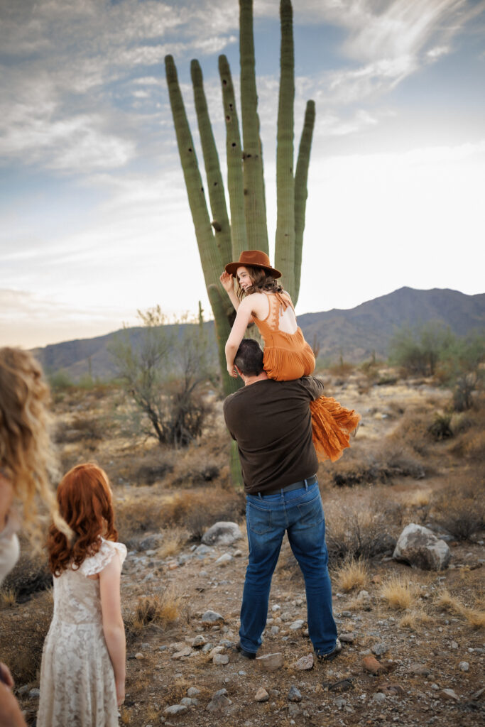 little girl in rust colored boho dress sitting on her dads shoulders with desert scenery in the background