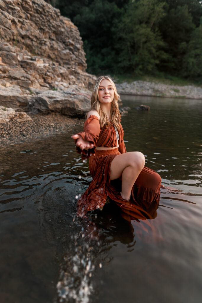 senior pictures in the water with girl splashing water toward the camera while wearing two piece boho gown