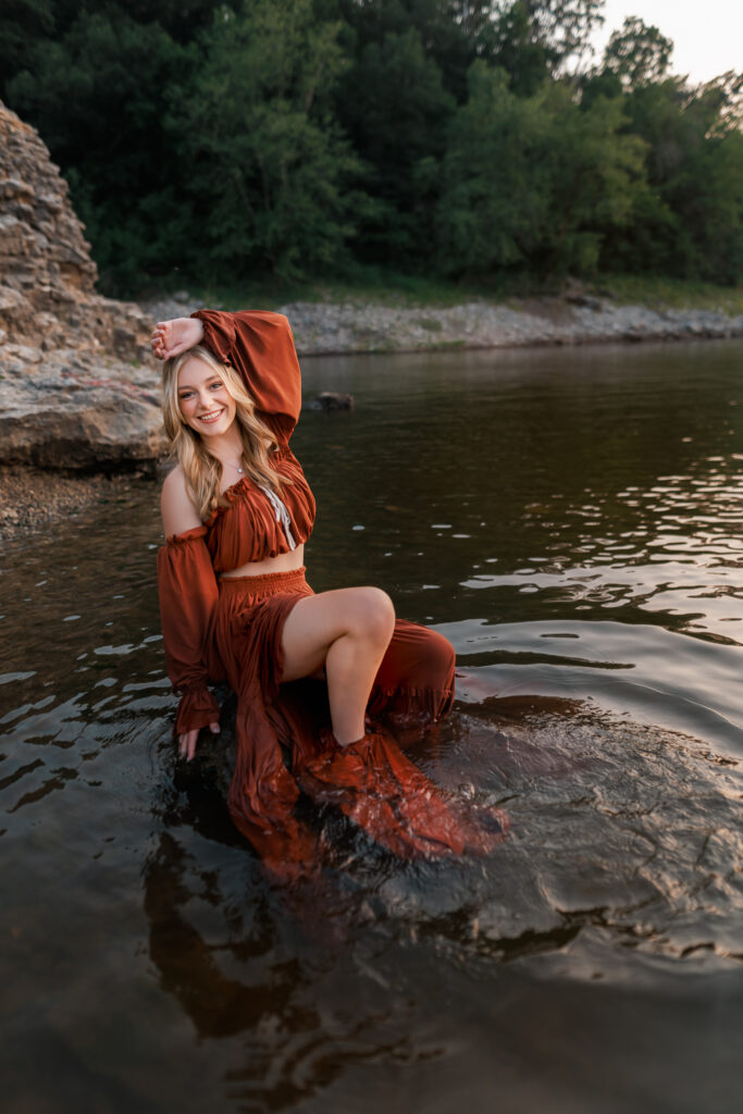 senior photos in the water with girl sitting on rock, arm above hear head, smiling at the camera