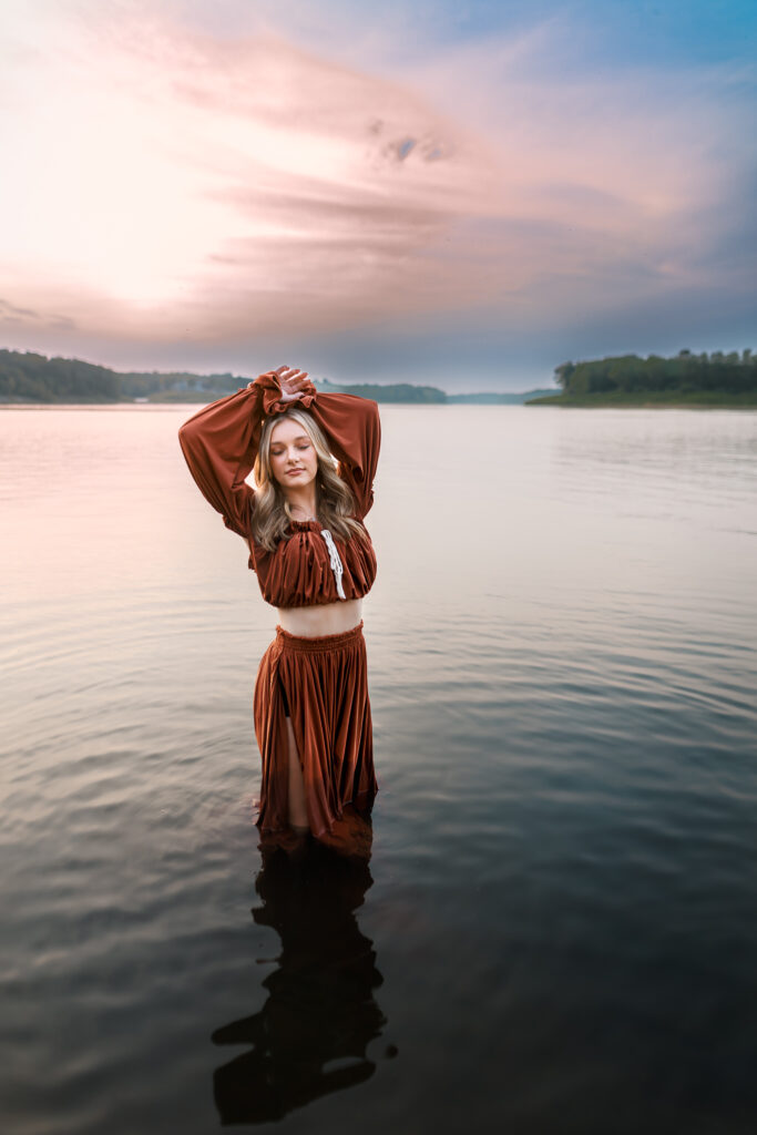 senior pictures in the water with girl holding hands above her head, eyes closed, and showing midriff with serene sunset in background