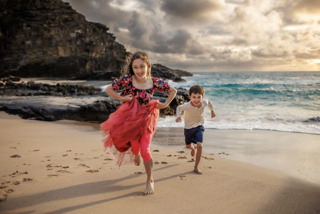 portrait of sibling running toward camera with water in background at beach photoshoot in hawaii