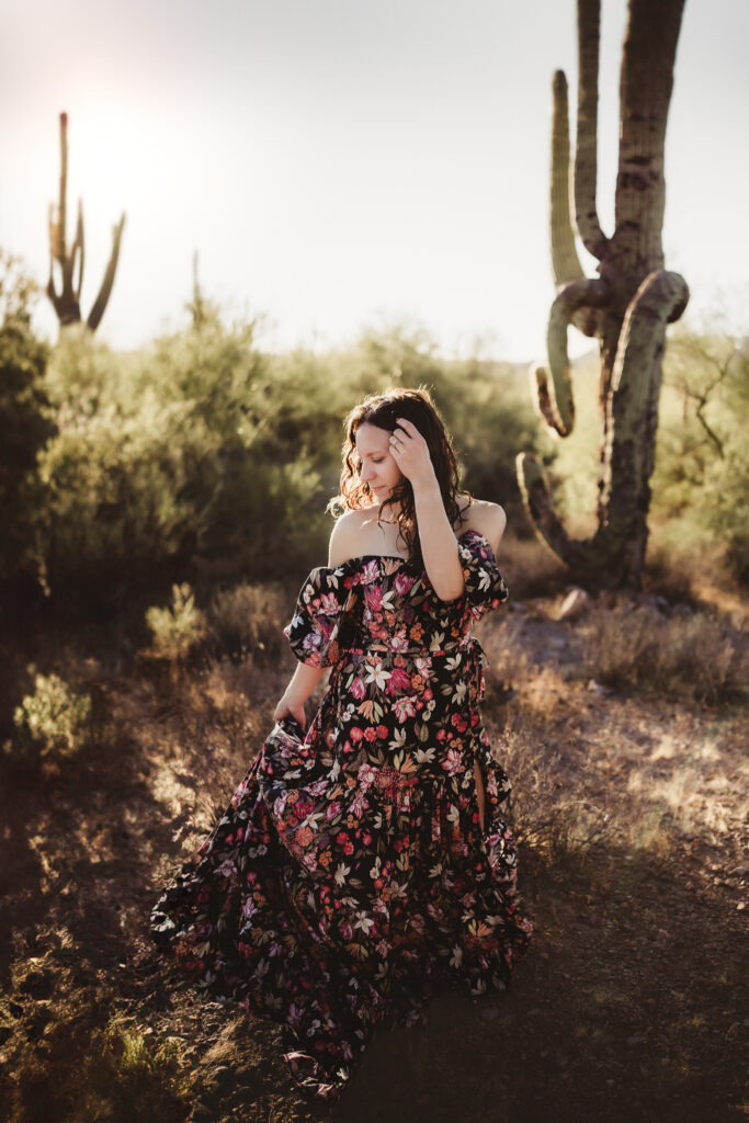 woman holding curly hair out of her face while standing in the desert in gloral gown near cactus